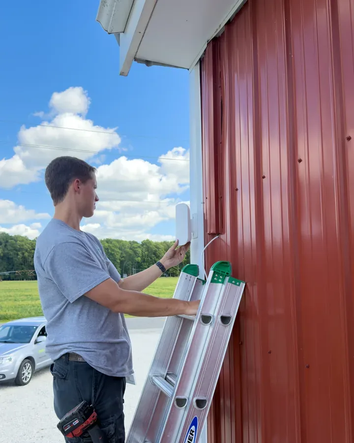 A Community Fiber technician on a ladder, mounting a fiber box on the side of a red metal building
